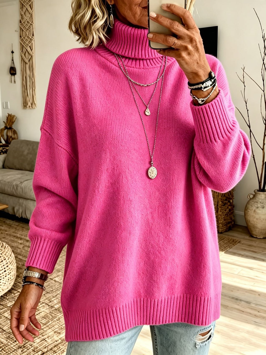 A woman takes a mirror selfie wearing an oversized bright pink turtleneck sweater, styled with layered silver necklaces and bracelets, in a cozy living room setting.