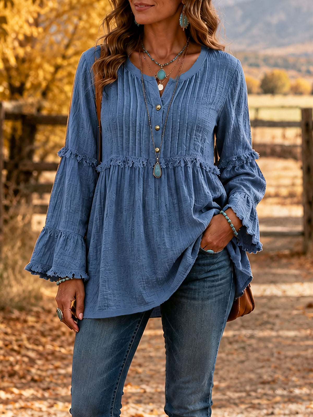 A woman wearing a blue long-sleeve blouse with a gathered peplum waist, ruffled cuffs, and front button details, paired with blue jeans in an outdoor autumn setting.