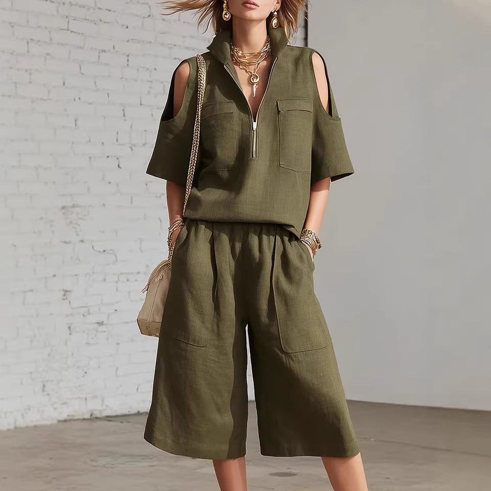 Woman wearing an olive green sleeveless top and shorts with a beige bag against a white brick wall.