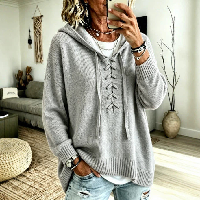 A woman takes a mirror selfie wearing a light gray oversized knit hoodie with a lace-up front, layered over a white top and paired with distressed light blue jeans in a cozy living room setting.