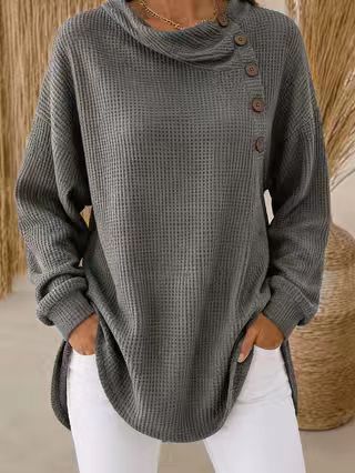 A woman wearing a dark gray waffle-knit cowl-neck sweater with brown buttons on the side, paired with white pants.