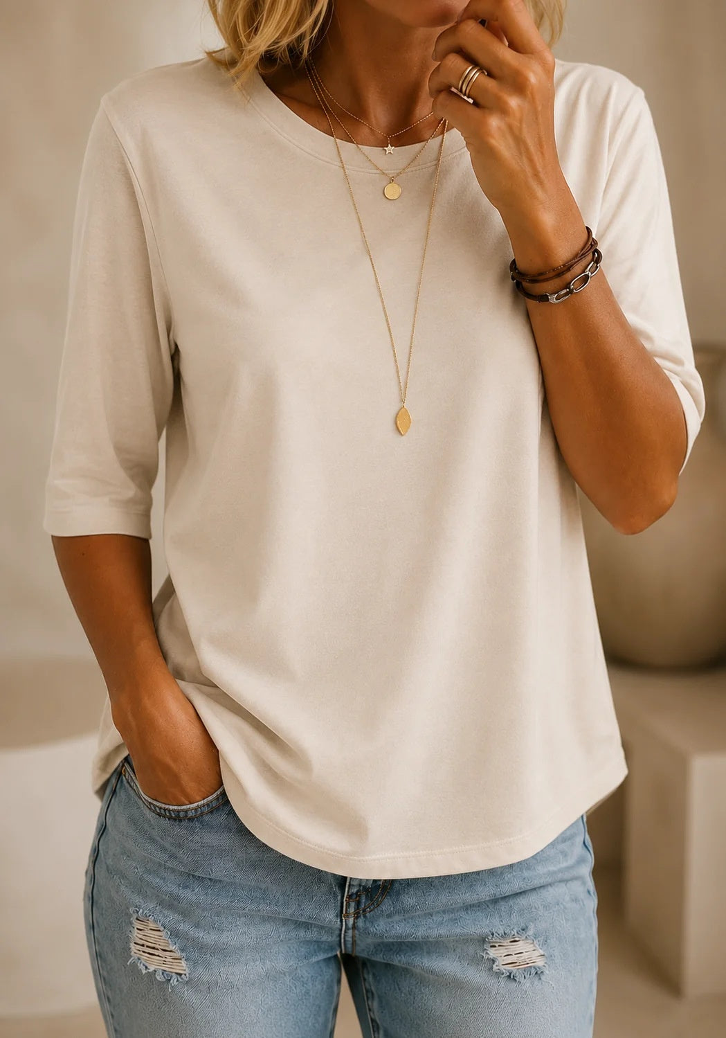 A woman wearing a cream-colored, relaxed-fit short-sleeve t-shirt and light-wash distressed blue jeans, accessorized with gold necklaces and bracelets.