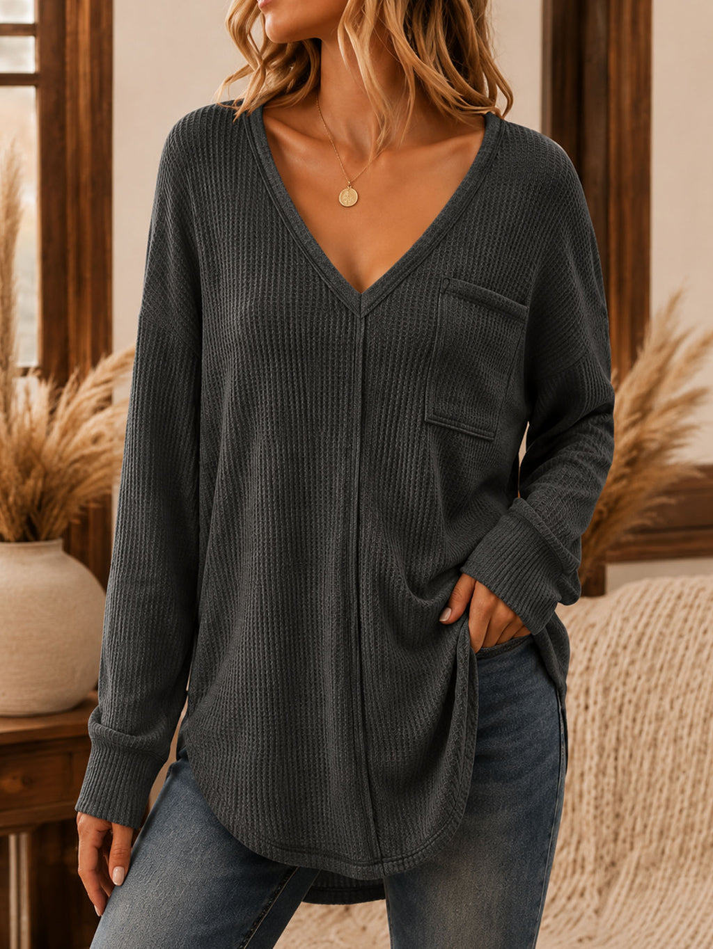 A woman wearing a dark charcoal gray ribbed knit long-sleeve top with a V-neck, button-front closure, and a chest pocket, paired with blue jeans in a warm indoor setting.