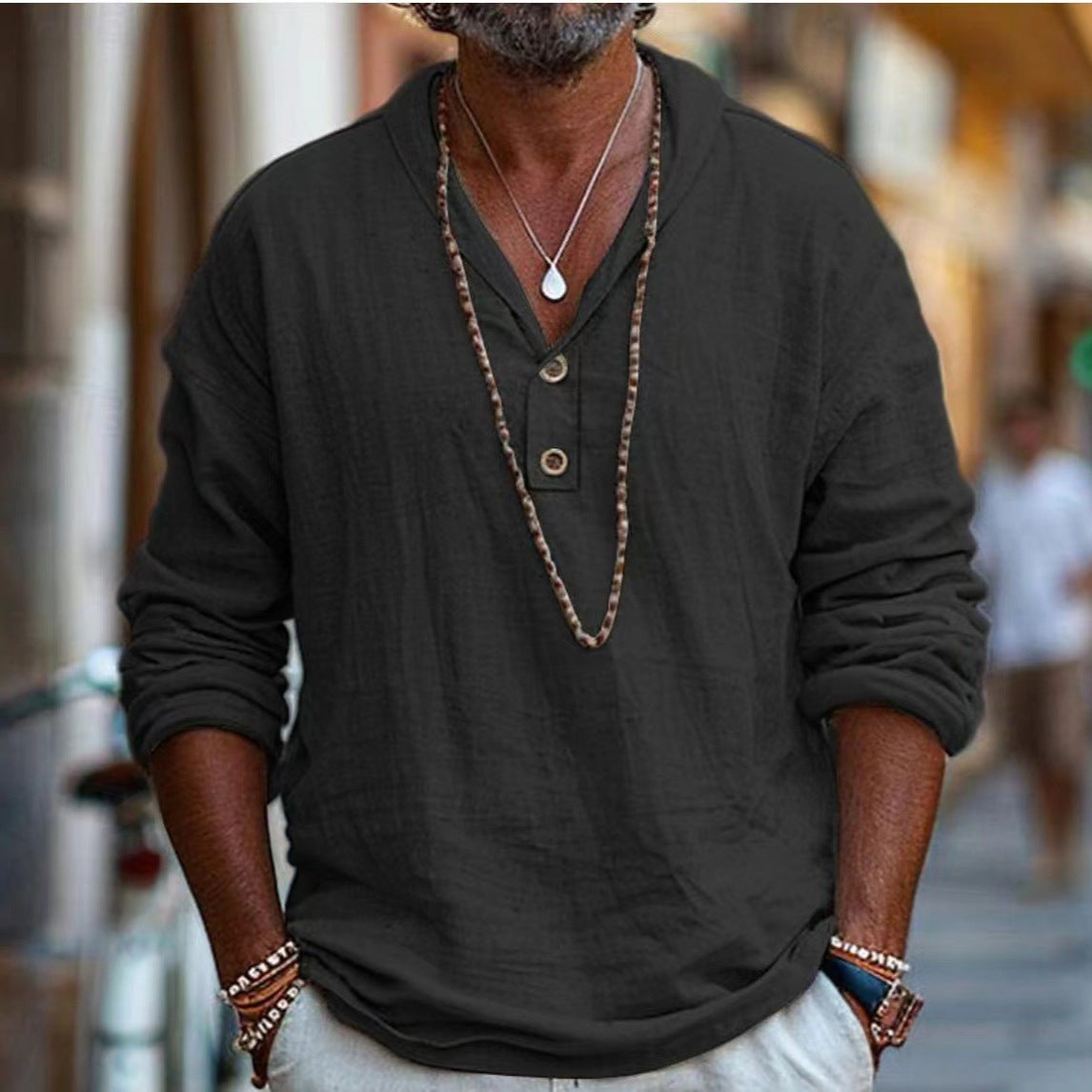 Man wearing a dark long-sleeve shirt with buttons, necklace, and bracelets in an urban setting.