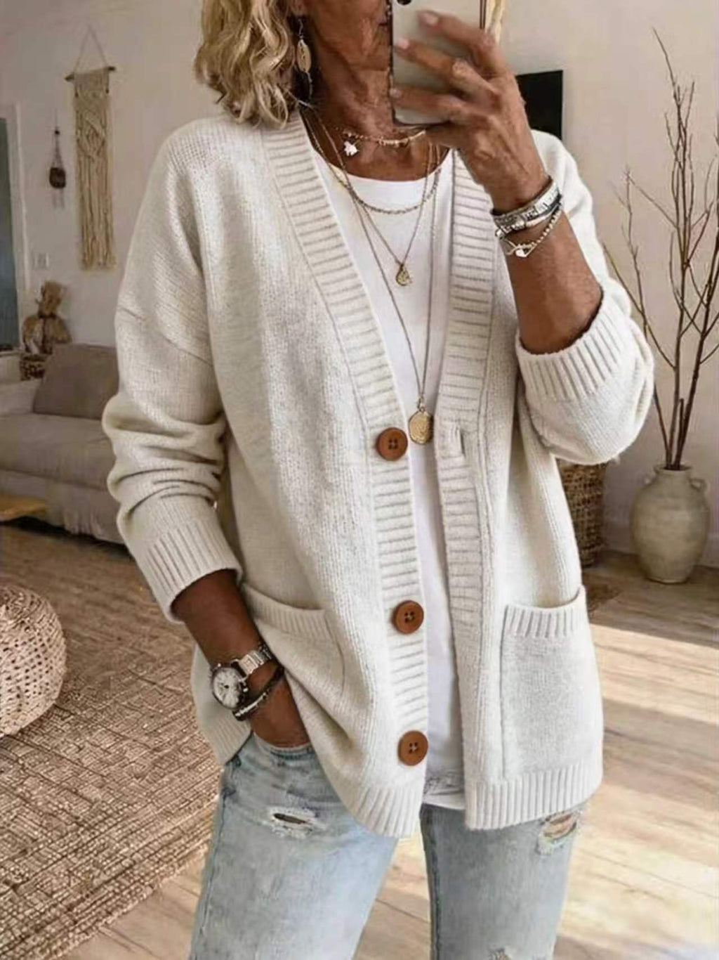 A woman takes a mirror selfie wearing a cream-colored knit cardigan with brown buttons, layered necklaces, and light-wash distressed jeans in a cozy living room setting.