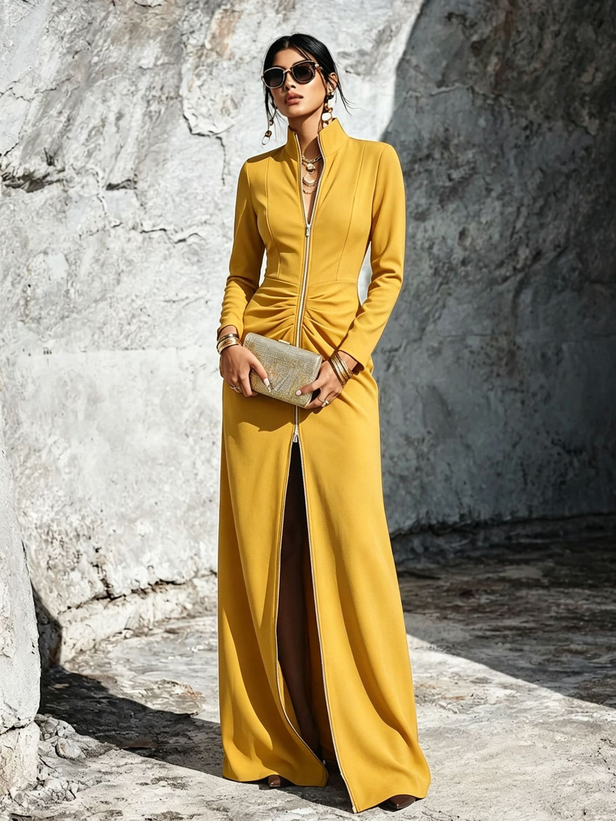 A woman stands outdoors against a textured stone wall wearing a long-sleeve mustard yellow maxi dress with a high collar, front zipper, and front slit. She is holding a metallic clutch and wearing sunglasses.