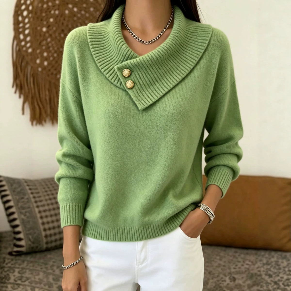 A woman wearing a light green knit sweater with a wide asymmetrical collar and two decorative gold-tone buttons, paired with white pants in a cozy indoor setting.