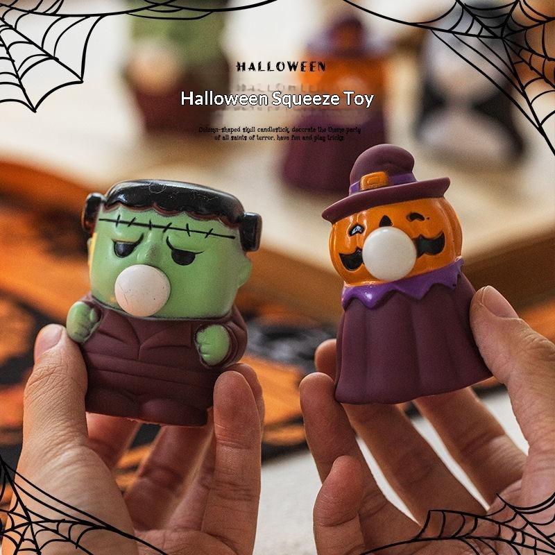 Two Halloween squeeze toys held in hands: a green Frankenstein figure with black hair and brown clothing, and an orange jack-o'-lantern figure wearing a purple witch hat and cape. The image has a blurred Halloween-themed background with spiderweb corner decorations and the text 'Halloween Squeeze Toy'.