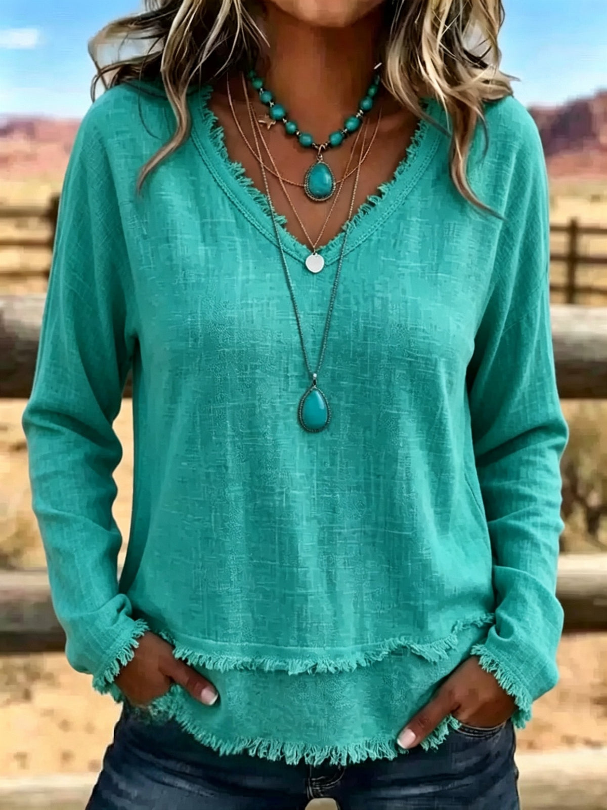A woman wearing a turquoise long-sleeve V-neck top with frayed edges, layered with turquoise and silver necklaces, standing outdoors in a desert landscape.
