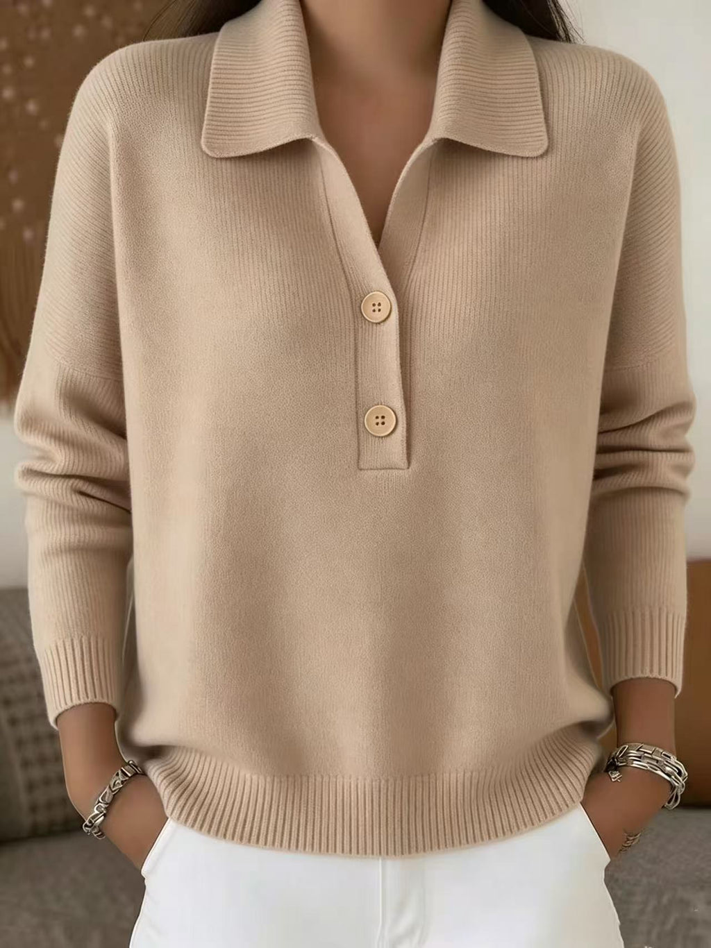 A woman wearing a beige knit polo sweater with three buttons and white pants, shown from the chest down in a softly lit indoor setting.