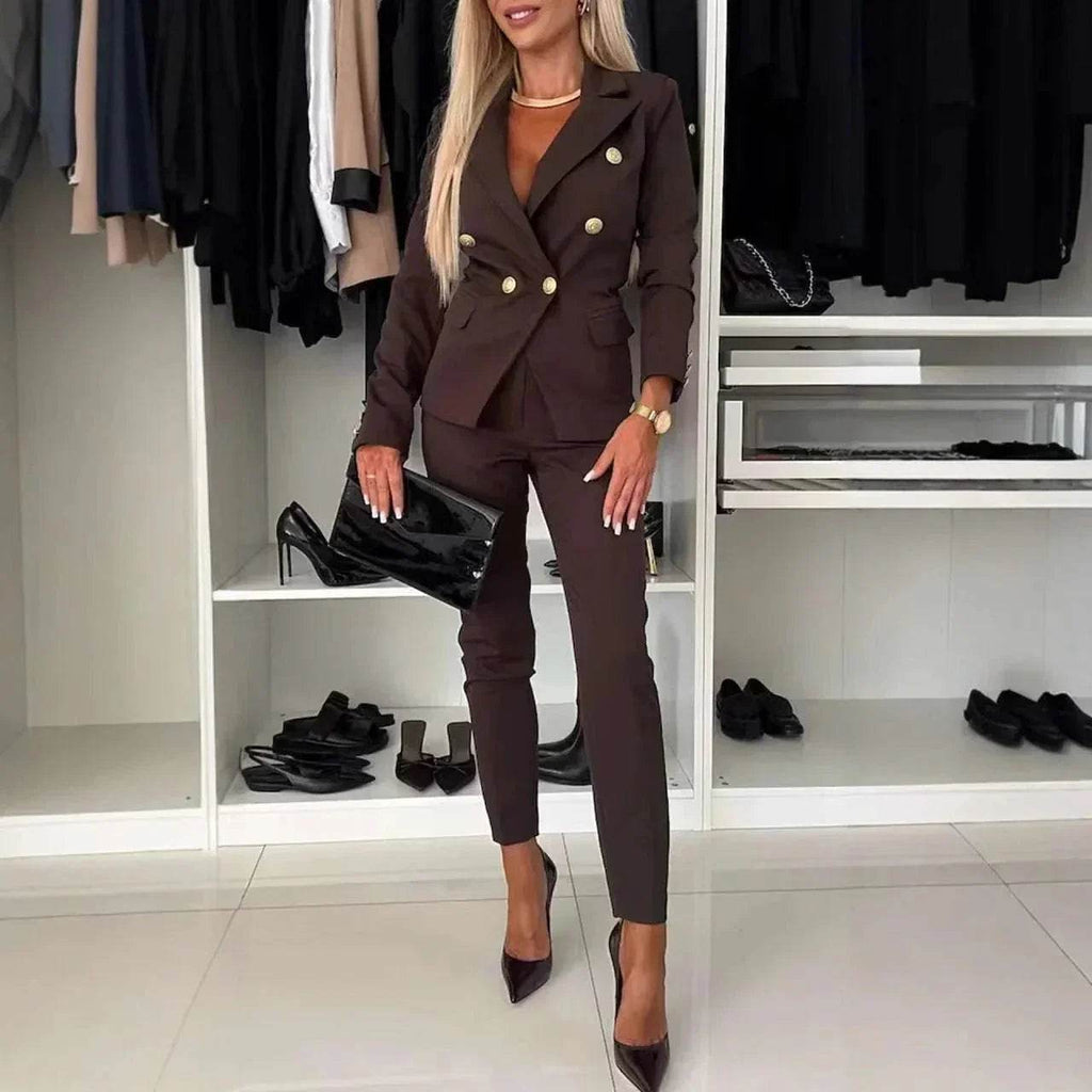 Woman in chic brown double-breasted pantsuit with clutch, posing in modern closet