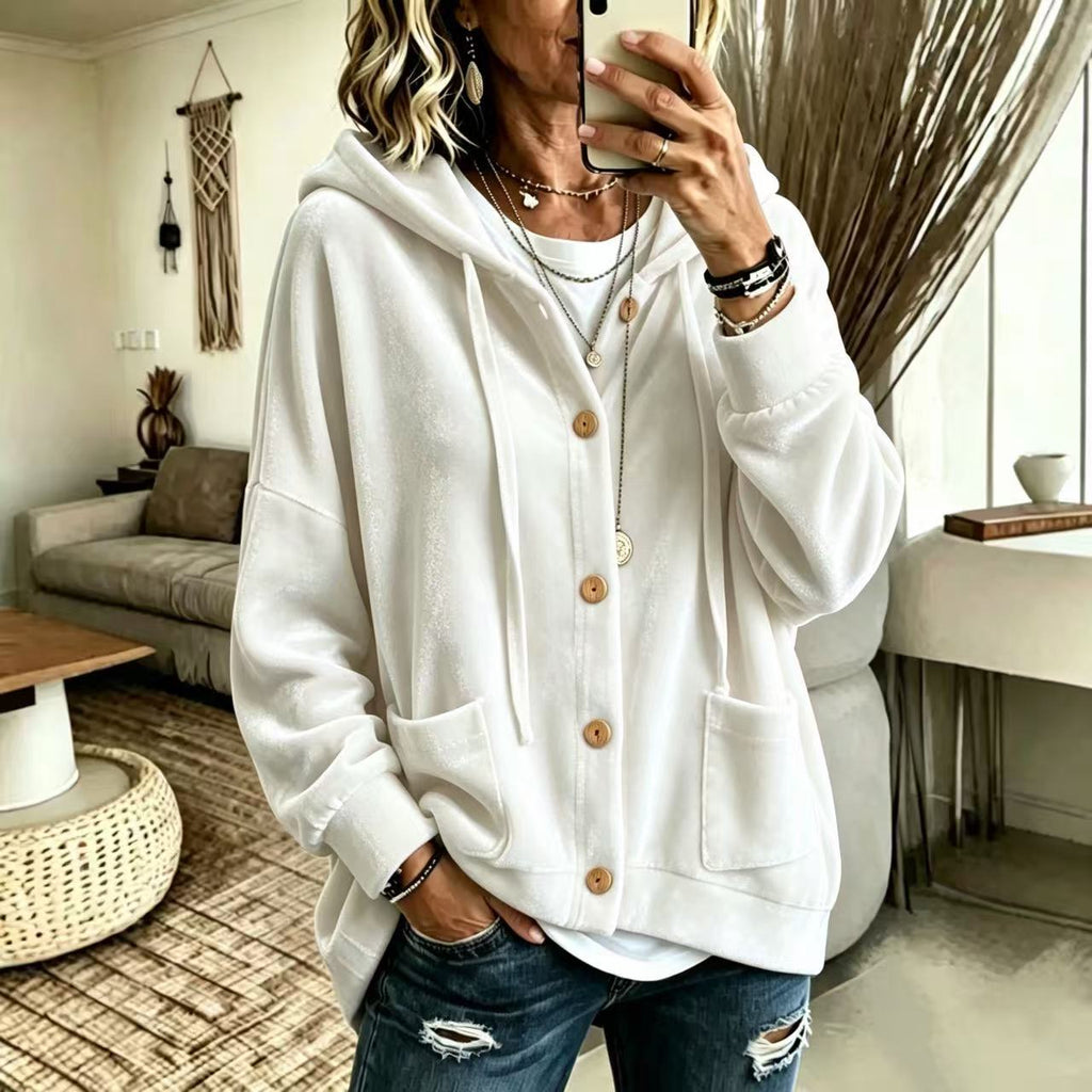A woman takes a mirror selfie wearing a cream-colored oversized button-up hoodie with a drawstring hood and front pockets, paired with distressed blue jeans in a modern living room setting.