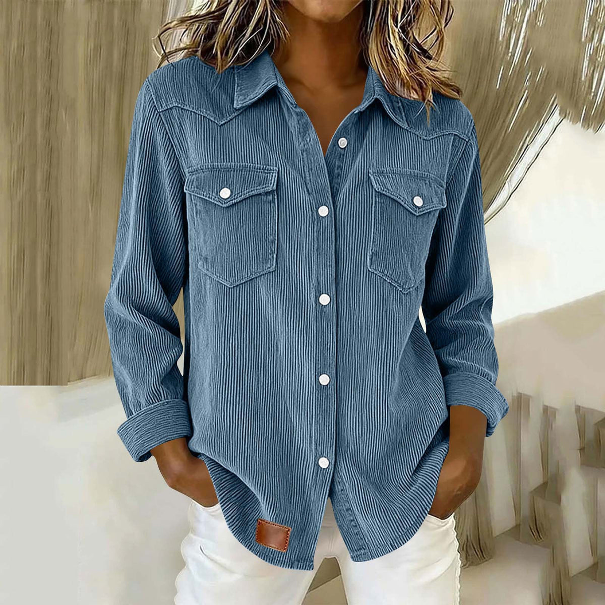 A woman wearing a blue corduroy button-up shirt with two chest pockets and white pants, standing indoors against a light neutral background.