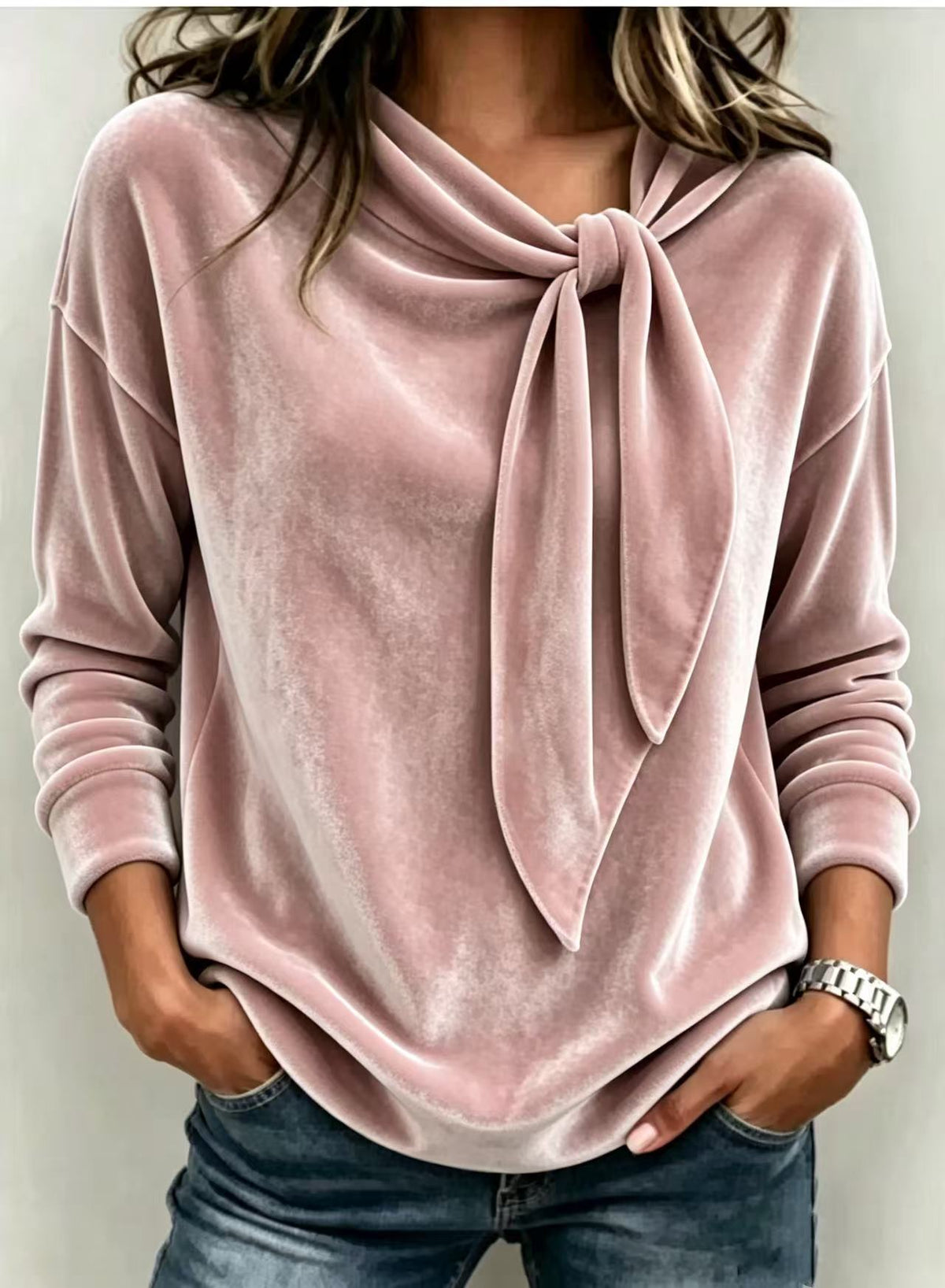 A woman wearing a dusty pink long-sleeve velvet top with a large front knot detail, paired with blue jeans.