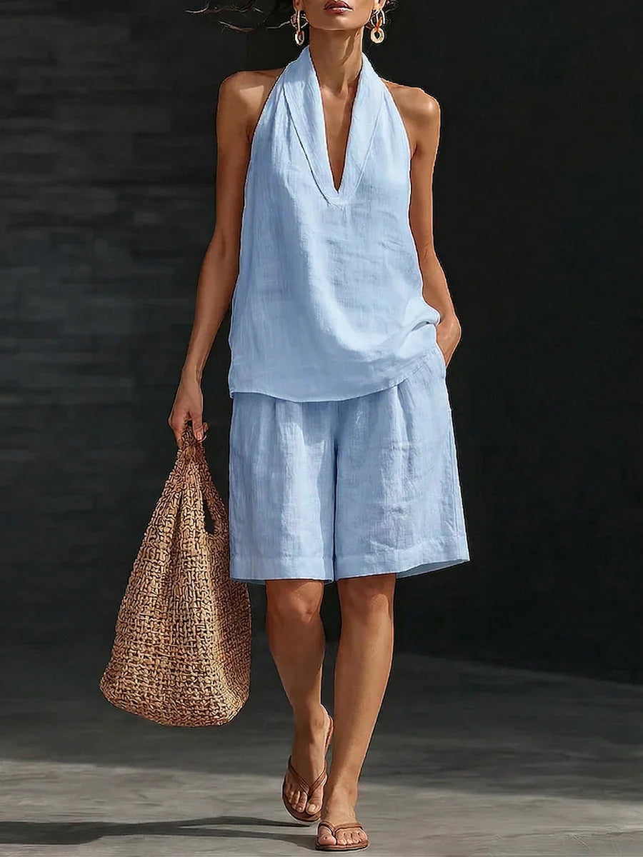 Woman wearing a light blue outfit with shorts and a top, holding a woven bag on a dark background.