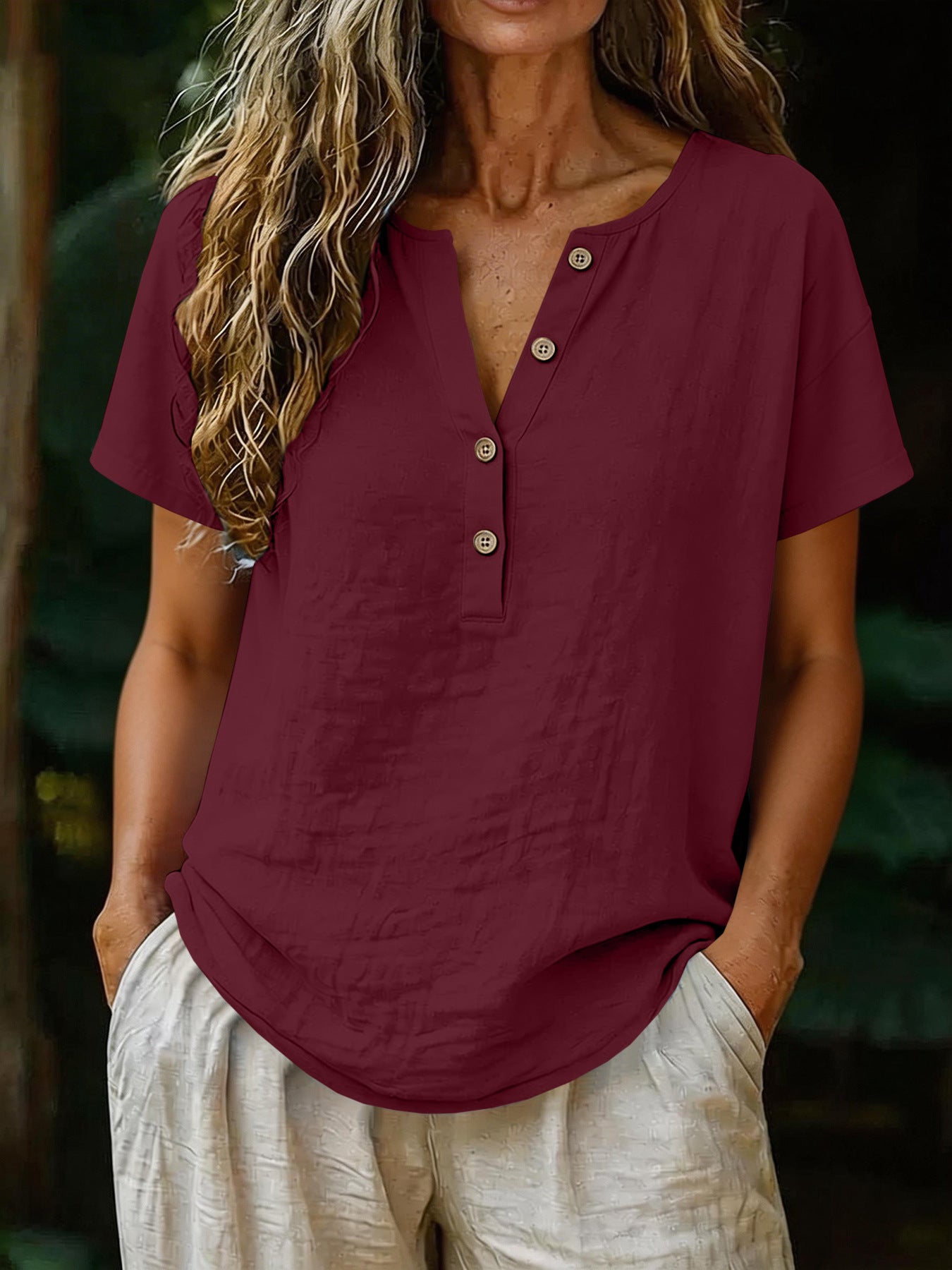 Person wearing a burgundy short-sleeve shirt with buttons, standing against a blurred natural background.