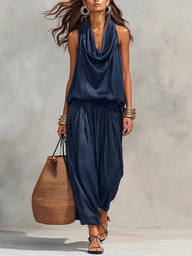 Woman wearing a navy blue sleeveless top and harem pants, holding a woven bag against a neutral background.