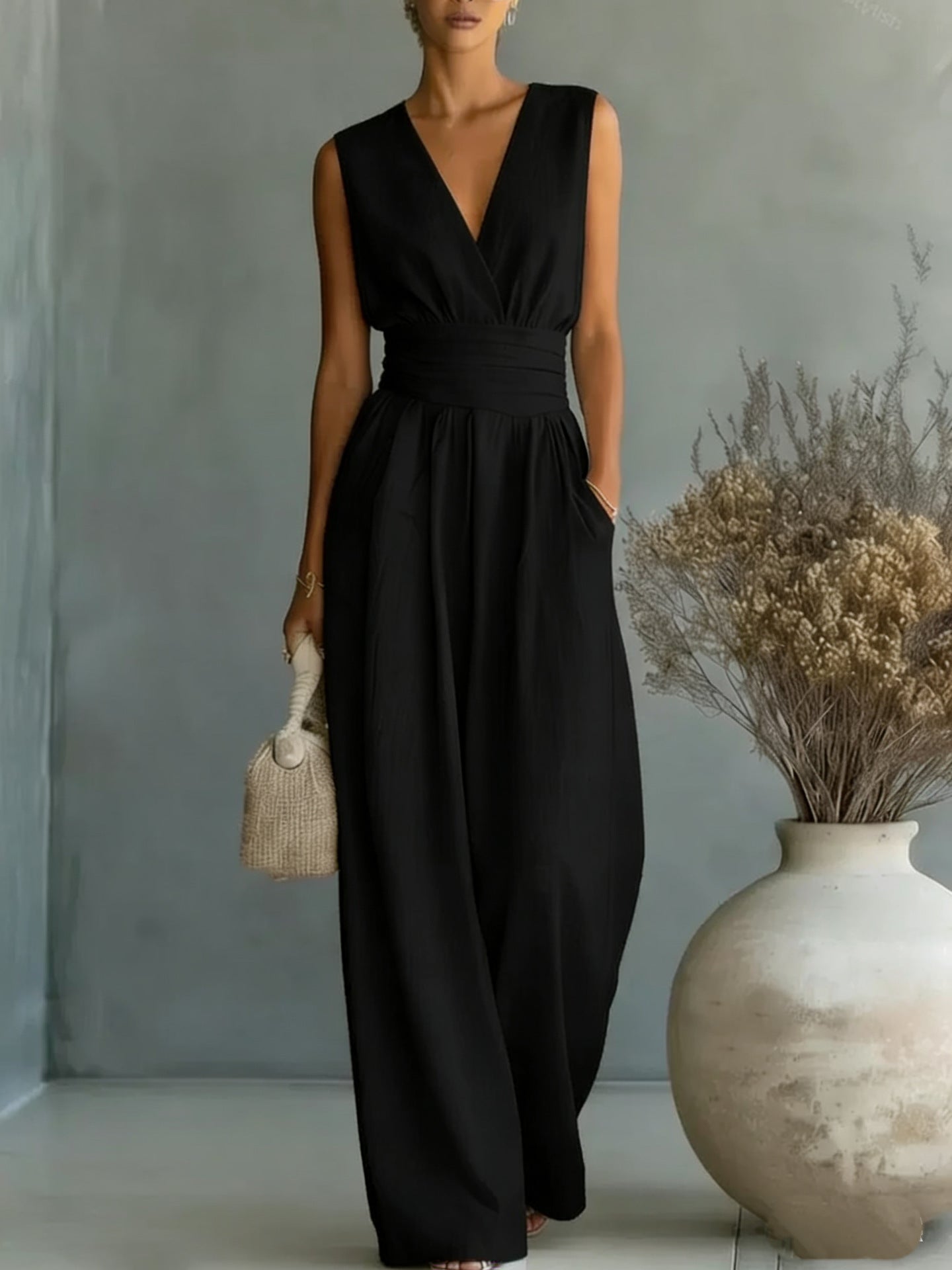 Woman wearing a black sleeveless jumpsuit holding a beige handbag against a neutral background.