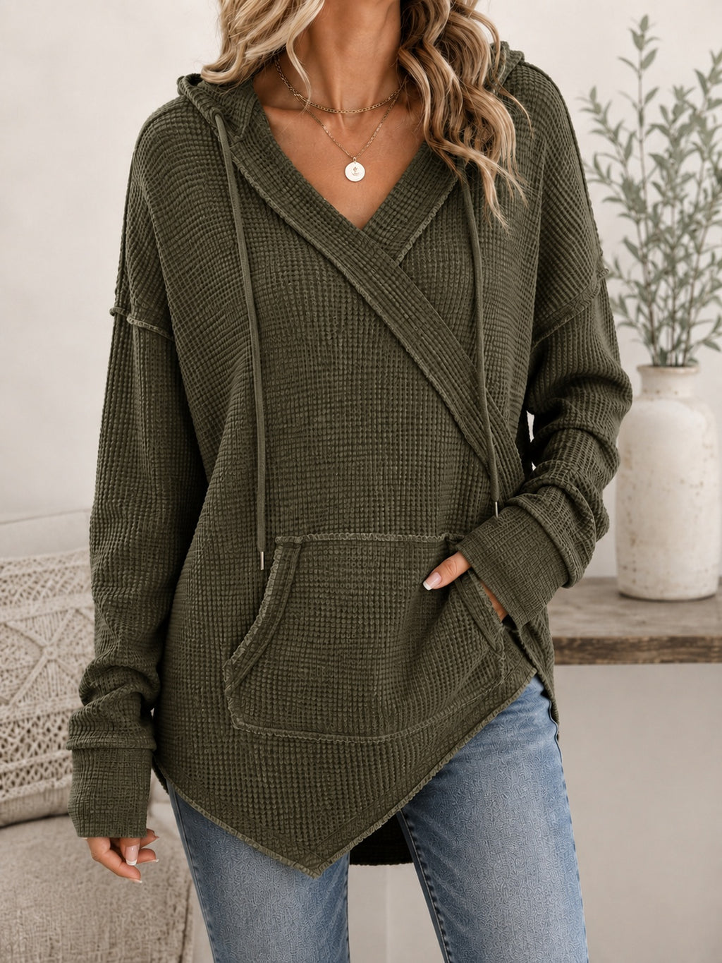 A woman wearing an olive green waffle-knit hooded sweater with a wrap-style front, drawstrings, and a kangaroo pocket, paired with blue jeans in a softly styled indoor setting.
