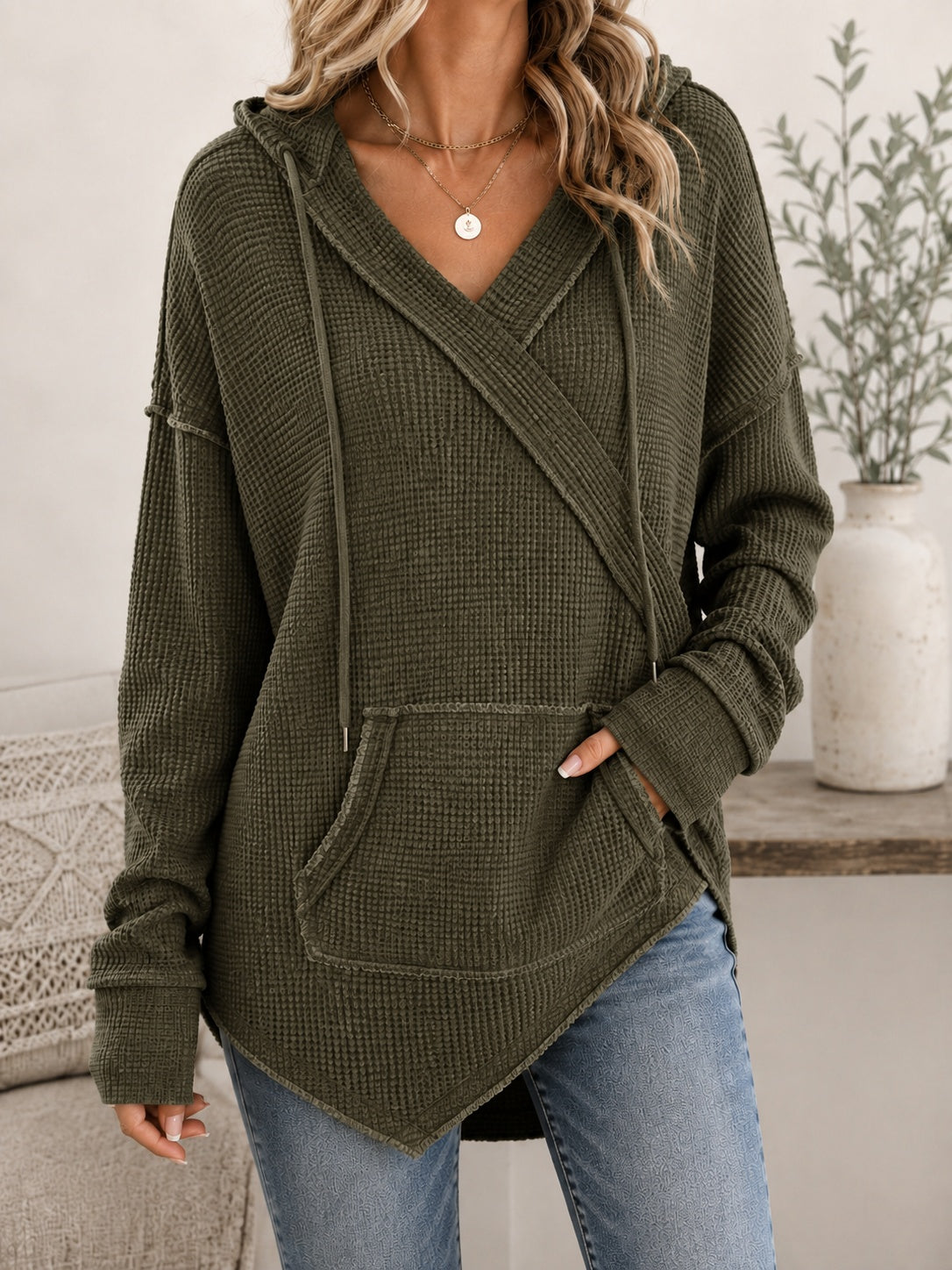 A woman wearing an olive green waffle-knit hooded sweater with a wrap-style front, drawstrings, and a kangaroo pocket, paired with blue jeans in a softly styled indoor setting.