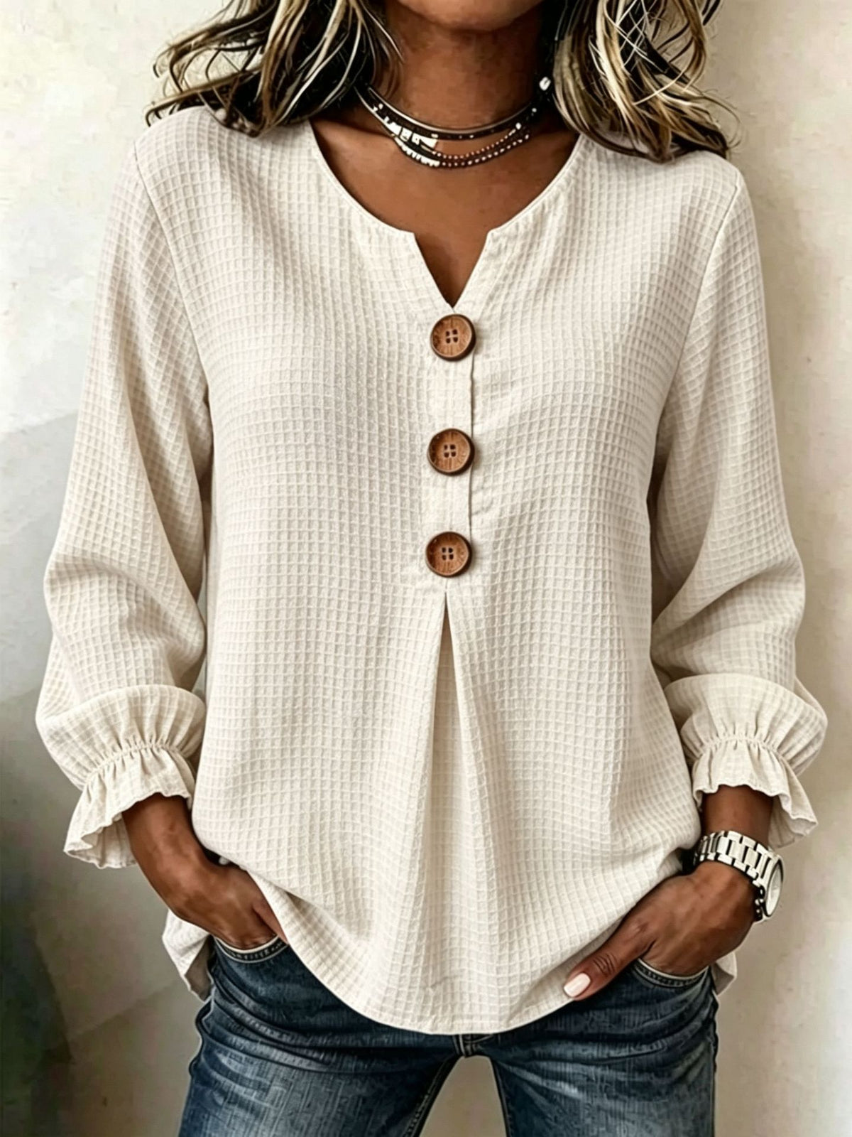 A woman wearing a cream-colored waffle-knit long-sleeve blouse with a split V-neck and three brown buttons, paired with blue jeans.