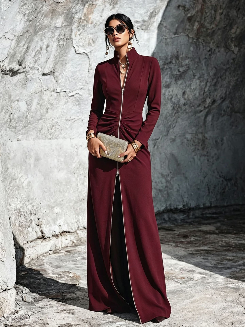 A woman models a long-sleeved, high-neck, floor-length maroon dress with a front zipper and a draped ruched waist, holding a metallic clutch against a textured stone wall.