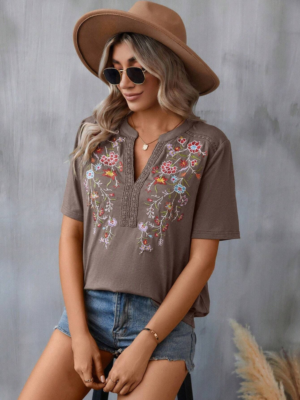 Woman wearing a brown embroidered shirt, denim shorts, and a brown hat against a gray background