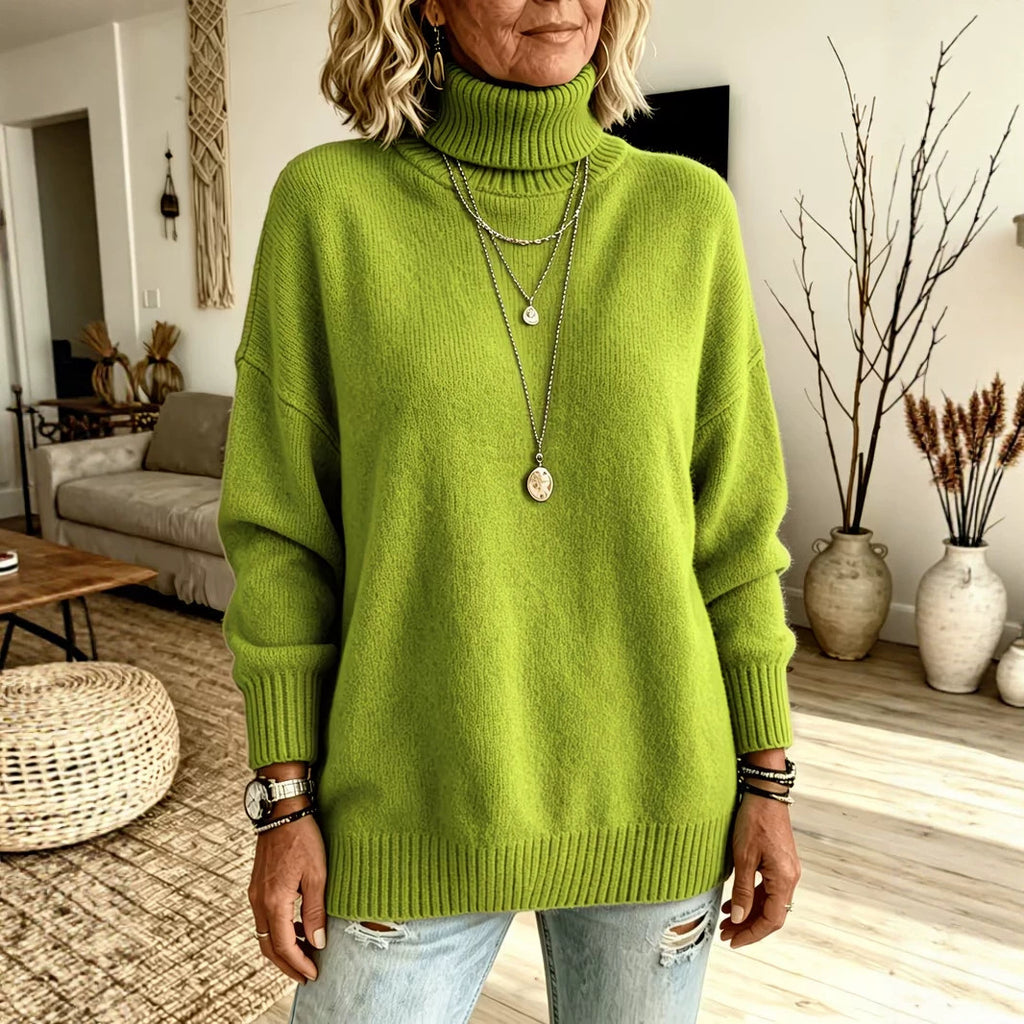 A woman wearing an oversized bright lime green turtleneck sweater, styled with layered silver necklaces and distressed light-wash jeans in a cozy, modern living room setting.