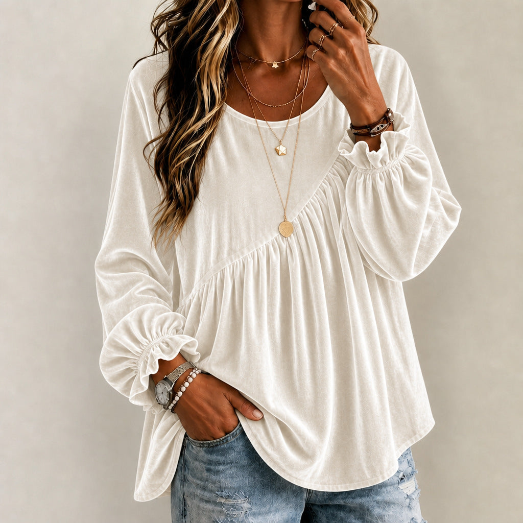 A woman wearing a white long-sleeve blouse with gathered ruffle details at the shoulders and cuffs, paired with light blue jeans and layered gold necklaces, against a plain light gray background.