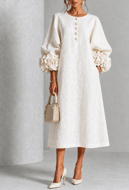A woman models a cream-colored midi dress with voluminous long sleeves, decorative ruffle cuffs, and a buttoned neckline. She is holding a small beige handbag and wearing white high heels, set against a neutral gray studio backdrop.