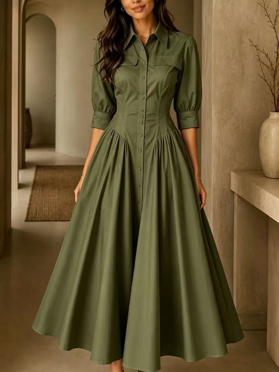 A woman wearing a long-sleeve olive green midi dress with a structured bodice, button front, and full flared skirt, standing in a warm-toned indoor hallway.