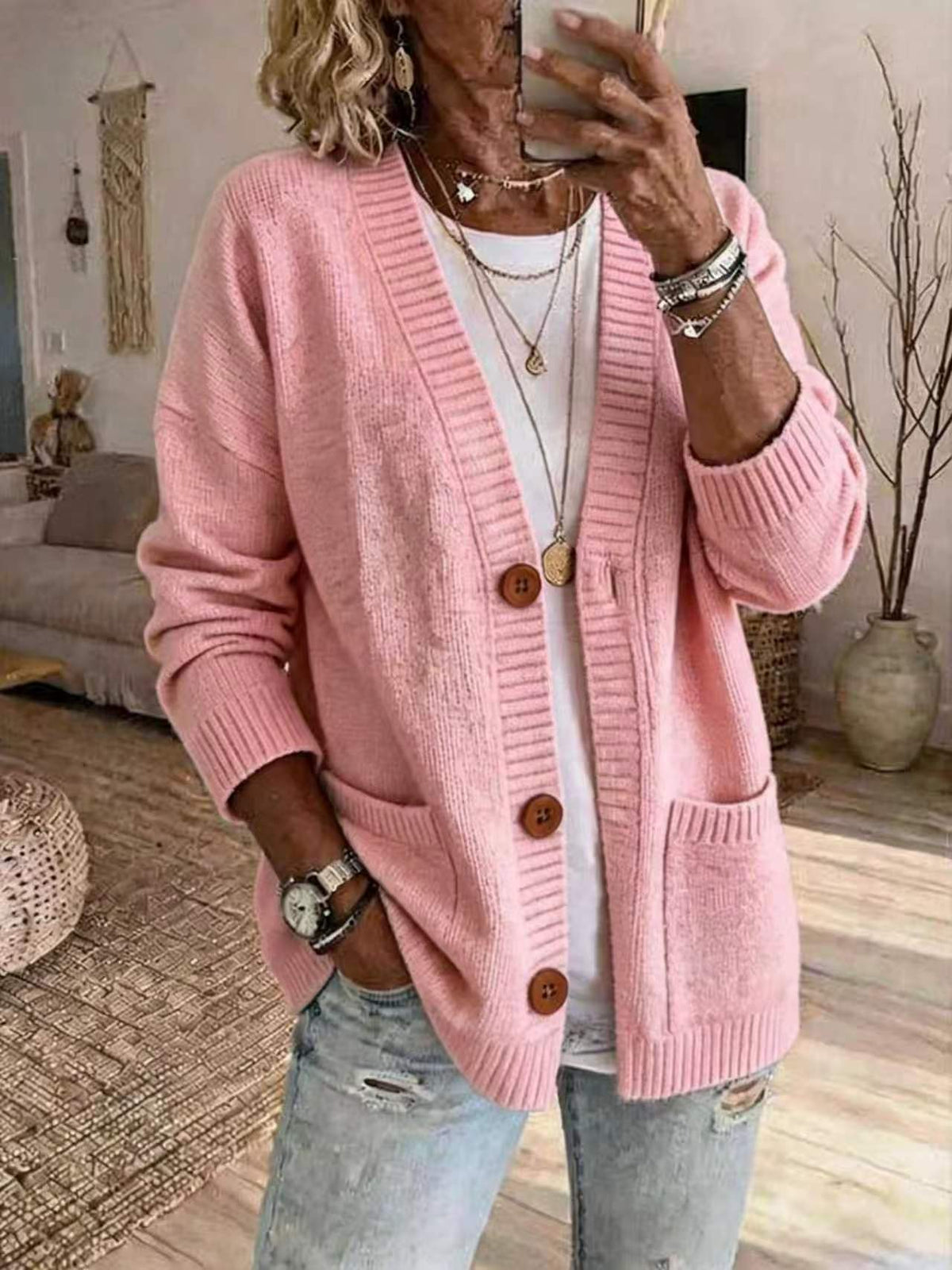 A woman wearing a pink open-front knit cardigan with brown buttons over a white top and light blue jeans, taking a mirror selfie in a living room setting.