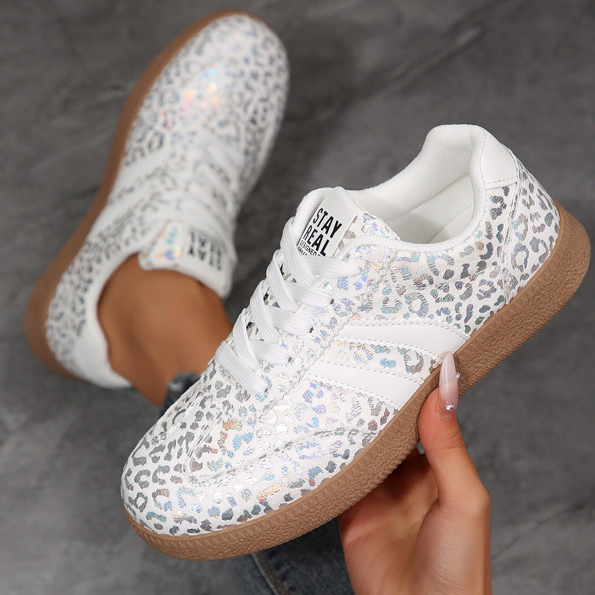 White sneakers with leopard print pattern and brown soles held by a hand on a gray background