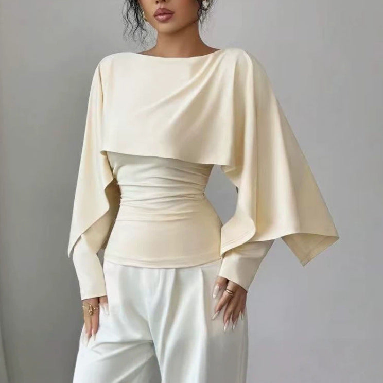A woman wearing a cream-colored draped long-sleeve top with a ruched waist and white pants, photographed against a plain gray studio background.