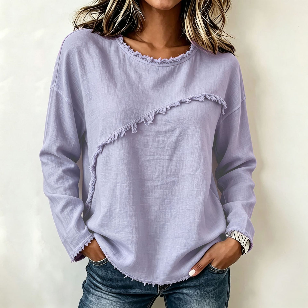 A woman wearing a light lavender long-sleeve top with a diagonal frayed detail and frayed hem, paired with blue jeans, against a plain off-white background.