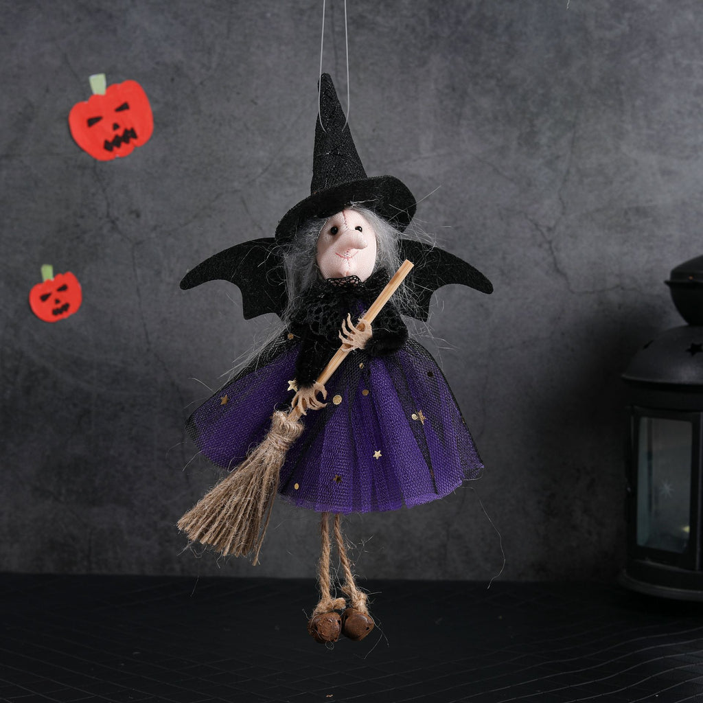 A hanging witch doll decoration with a black pointed hat, gray hair, a purple skirt with gold stars, and a straw broom, photographed against a dark textured wall with small pumpkin props.