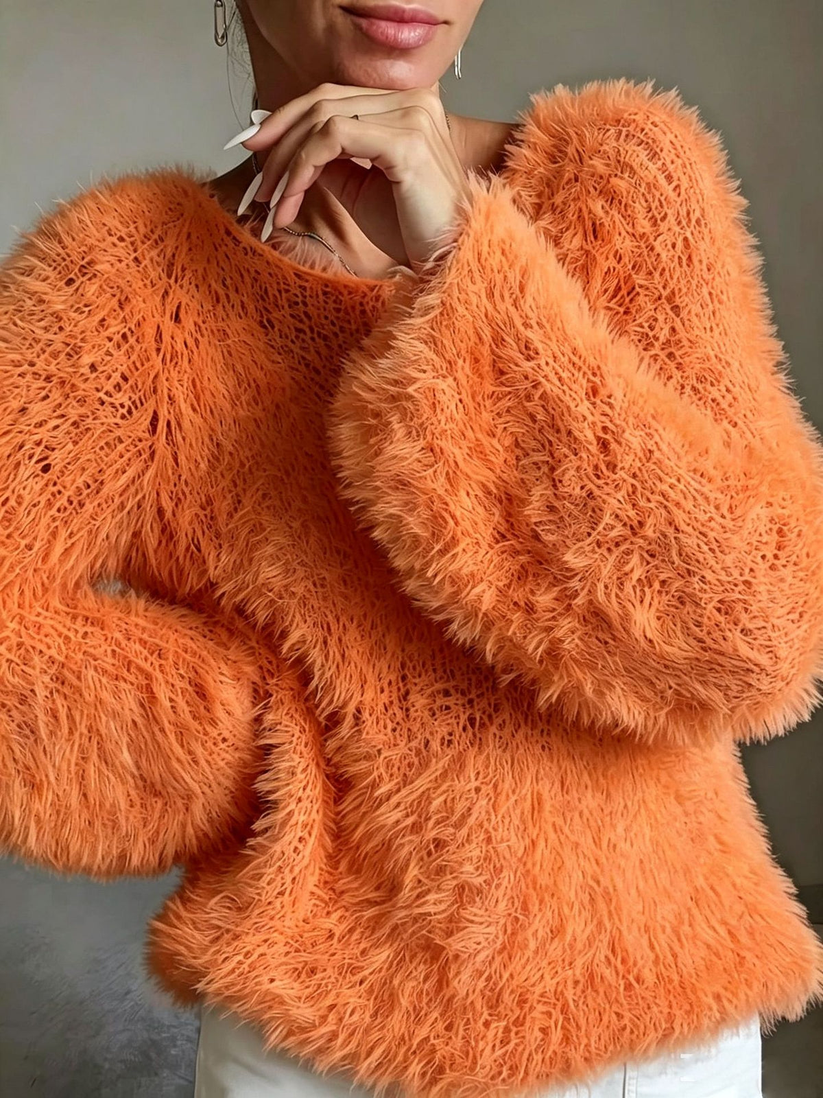 A close-up of a person wearing a fuzzy, textured orange knit sweater with voluminous long sleeves and a crew neckline, paired with white pants.