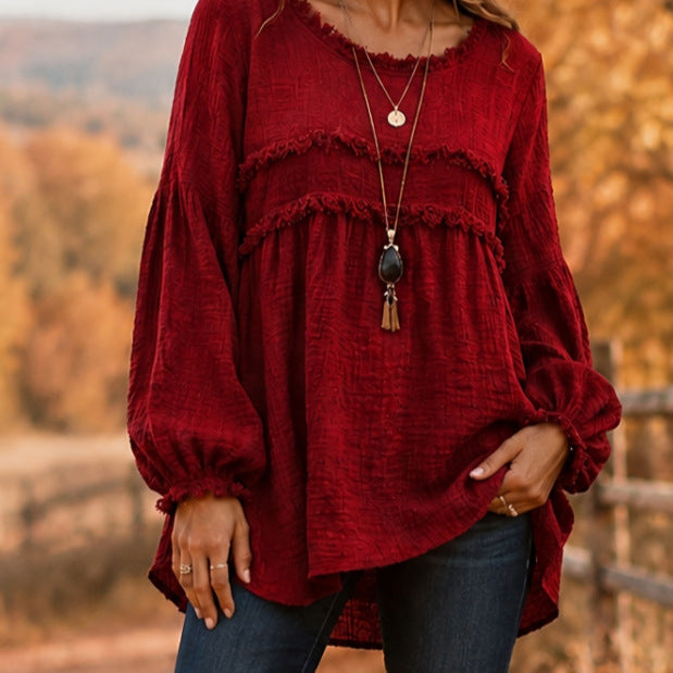 A woman wearing a long-sleeve, deep burgundy red blouse with layered ruffle details and a relaxed fit, paired with blue jeans and layered necklaces, set against a blurred autumn outdoor background.