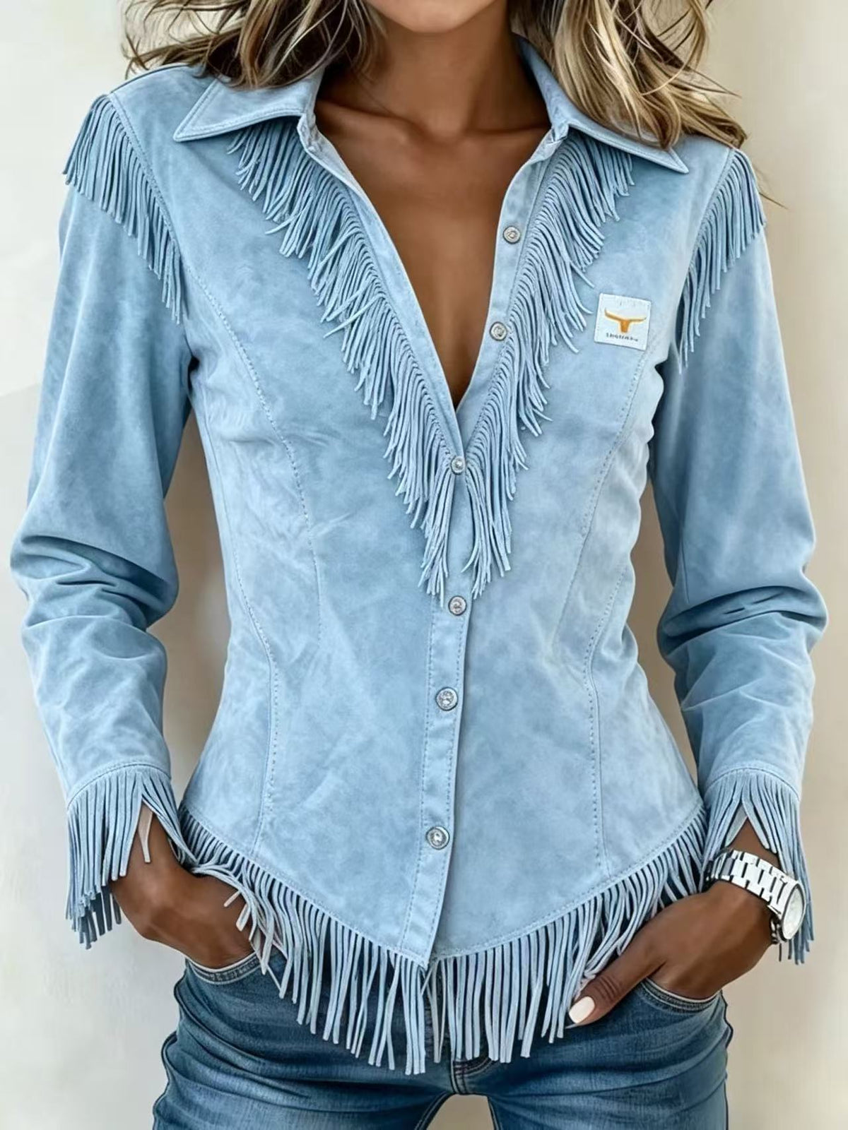 A woman wearing a light blue suede long-sleeve western-style shirt with fringe detailing and a small Texas Longhorns logo patch on the chest, paired with blue jeans.