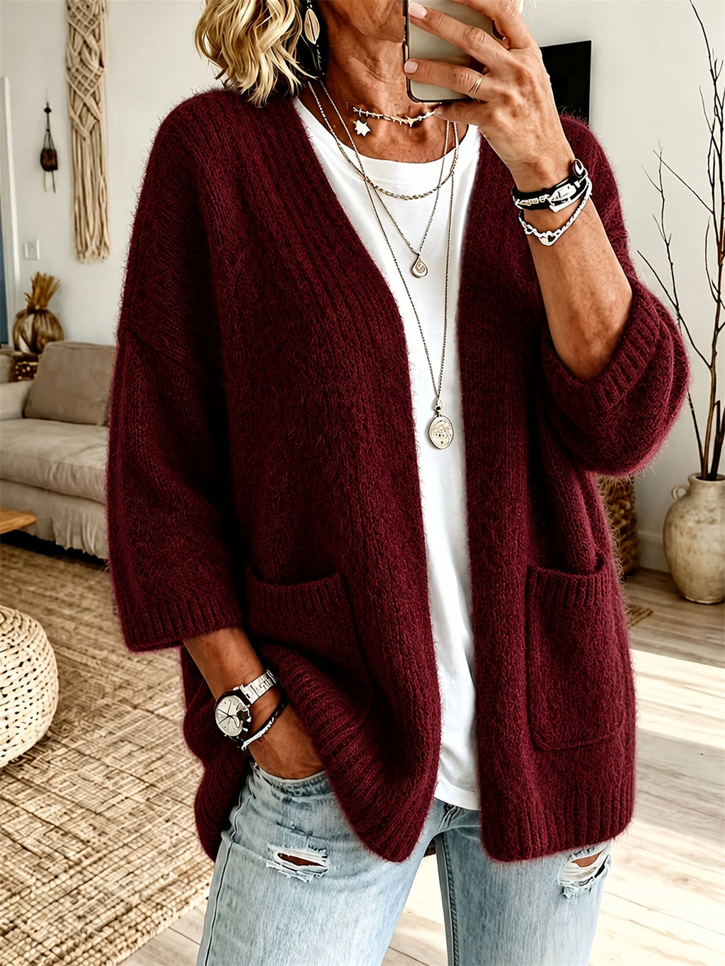 A woman wearing a chunky knit open-front cardigan in deep burgundy over a white t-shirt and light-wash distressed jeans, photographed in a cozy living room setting.