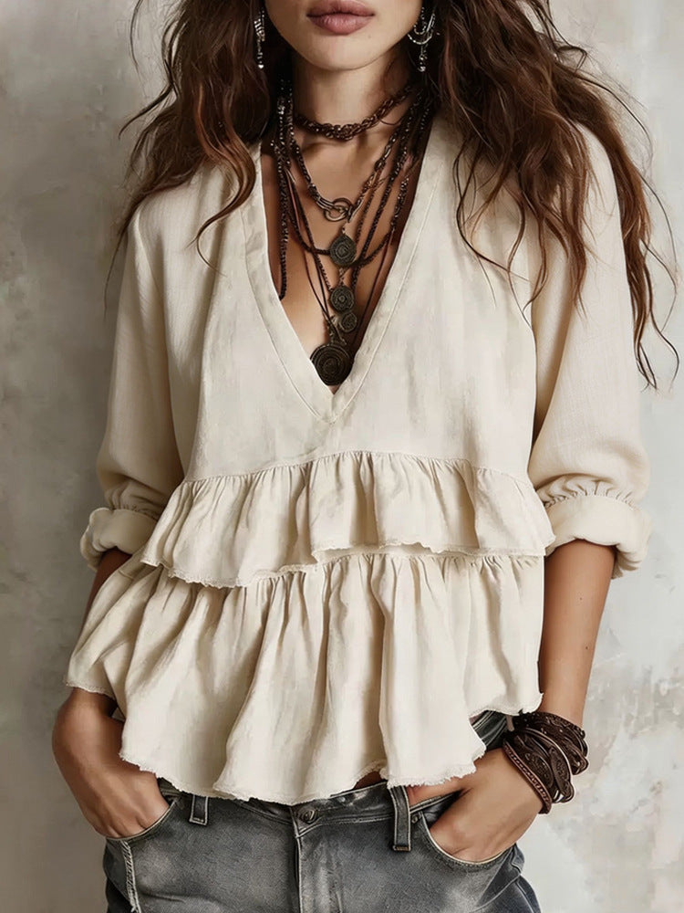 A woman wearing a cream-colored, long-sleeve bohemian-style blouse with a deep V-neck and tiered ruffle details, paired with layered dark brown necklaces and bracelets, set against a textured gray wall.