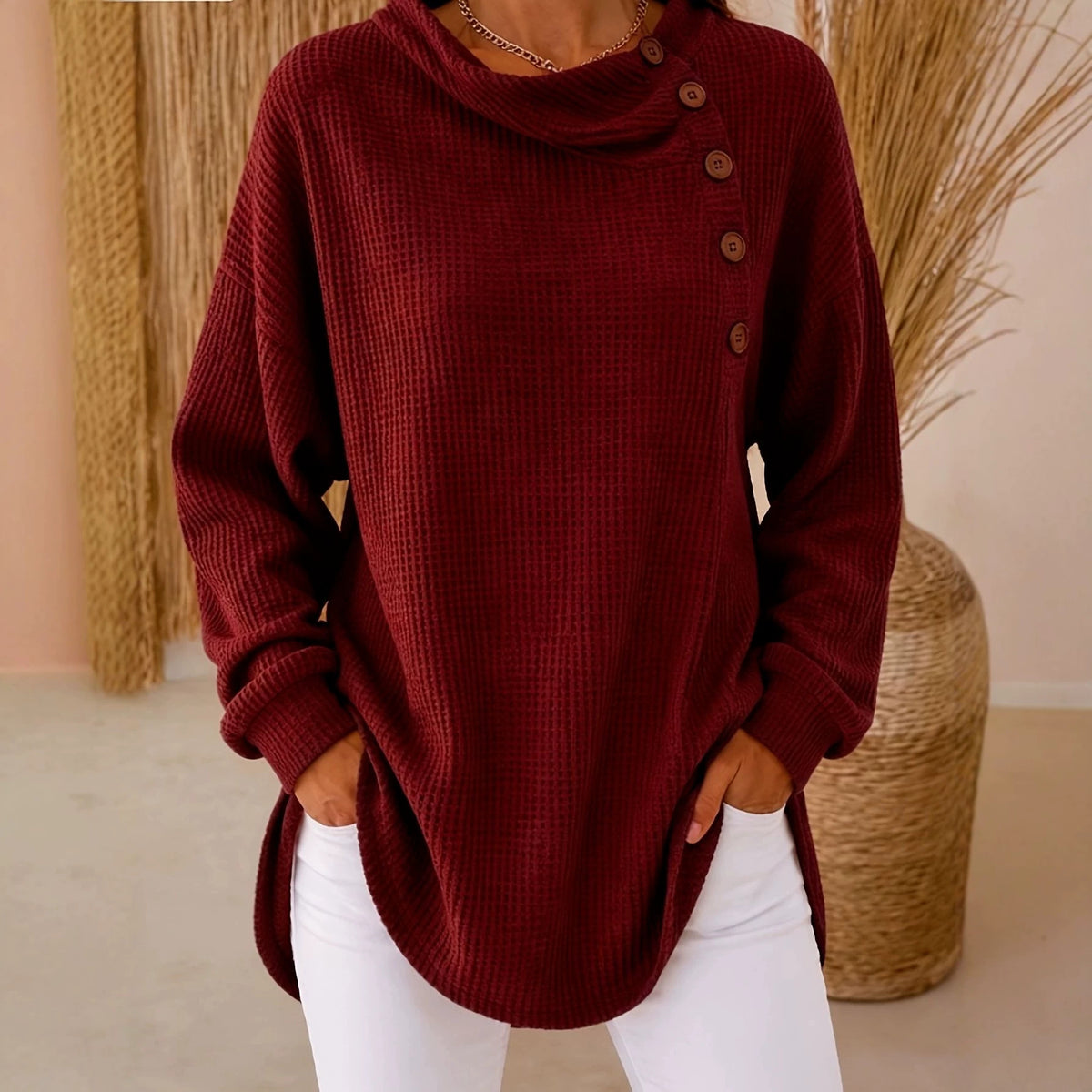 A woman wearing a long-sleeve waffle-knit cowl-neck sweater in deep burgundy with brown buttons down the shoulder, paired with white pants, standing in a styled indoor setting with dried grass decor.