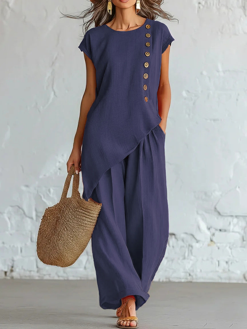 Woman wearing a long navy dress with button details, holding a woven bag against a white brick wall.