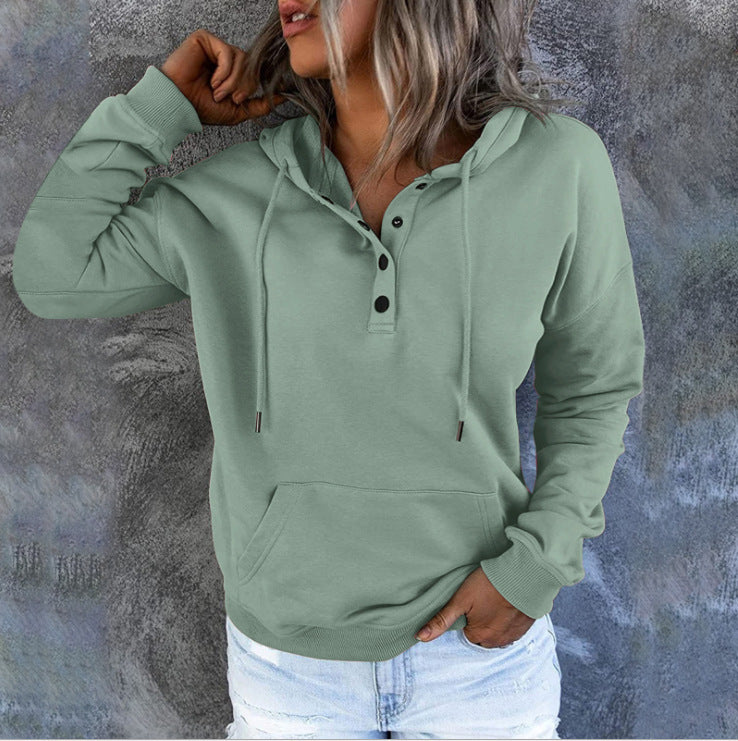 A woman wearing a sage green pullover hoodie with a buttoned henley-style collar, drawstrings, and a front kangaroo pocket, paired with light blue jeans, set against a textured gray wall.