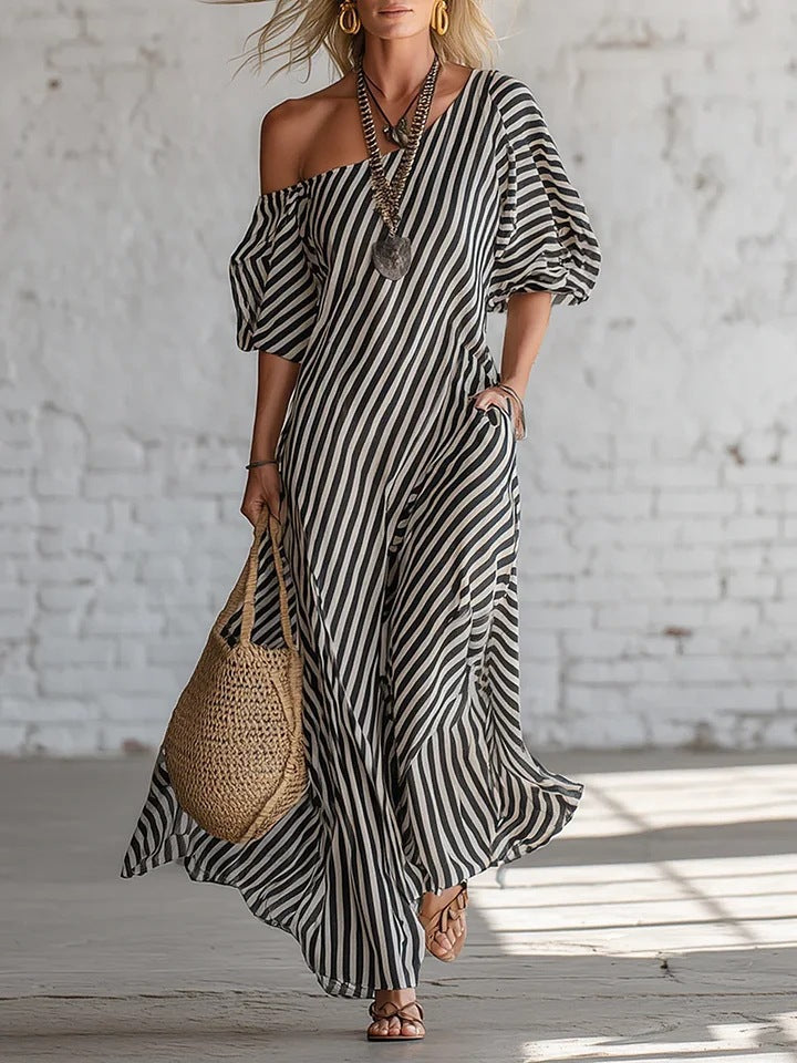 Woman wearing a black and white striped dress with a woven bag against a white brick wall.