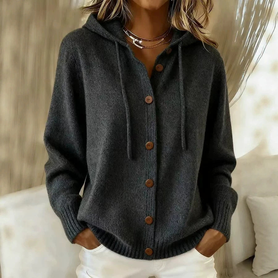 A woman wearing a dark gray knit hooded cardigan with brown buttons, paired with white pants, in a softly lit indoor setting.