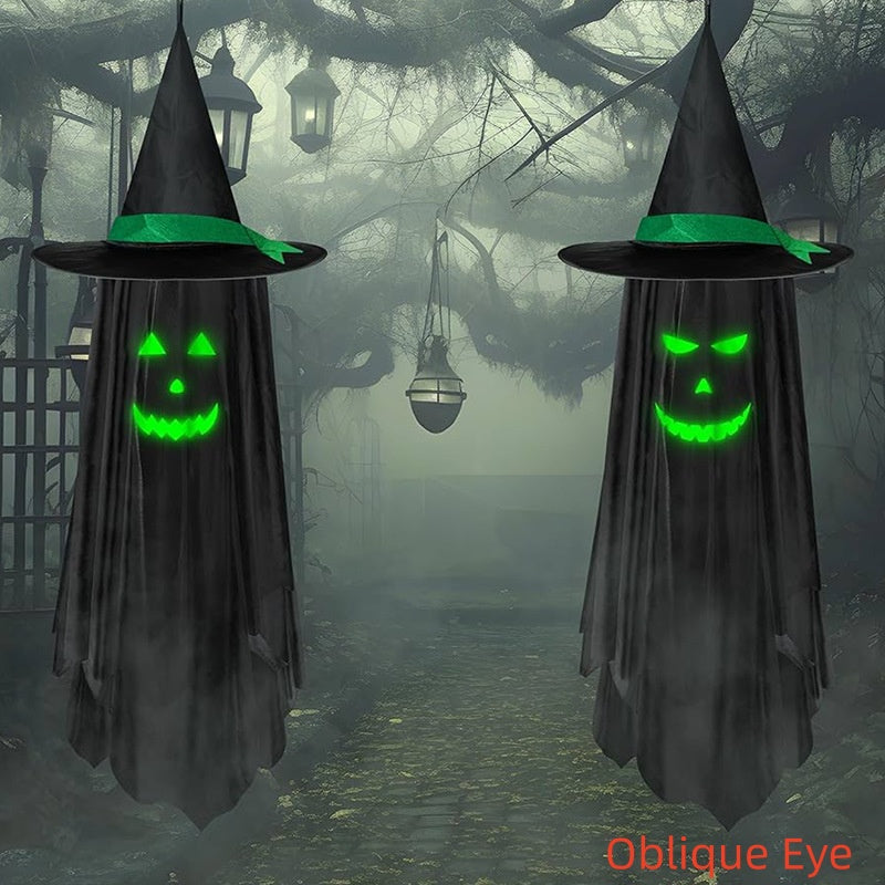 Two hanging black witch cloaks with pointed hats and glowing green jack-o'-lantern faces, set against a foggy Halloween forest path with lanterns and bare trees. The text 'Oblique Eye' appears in orange at the bottom right.