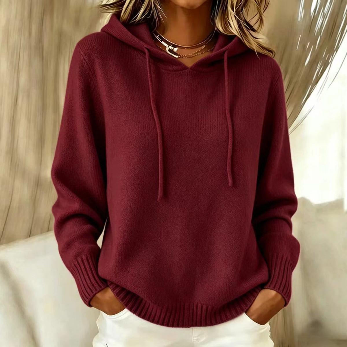 A woman wearing a ribbed knit pullover hoodie in deep burgundy with matching drawstrings, paired with white pants and layered gold necklaces, set against a softly lit indoor background.