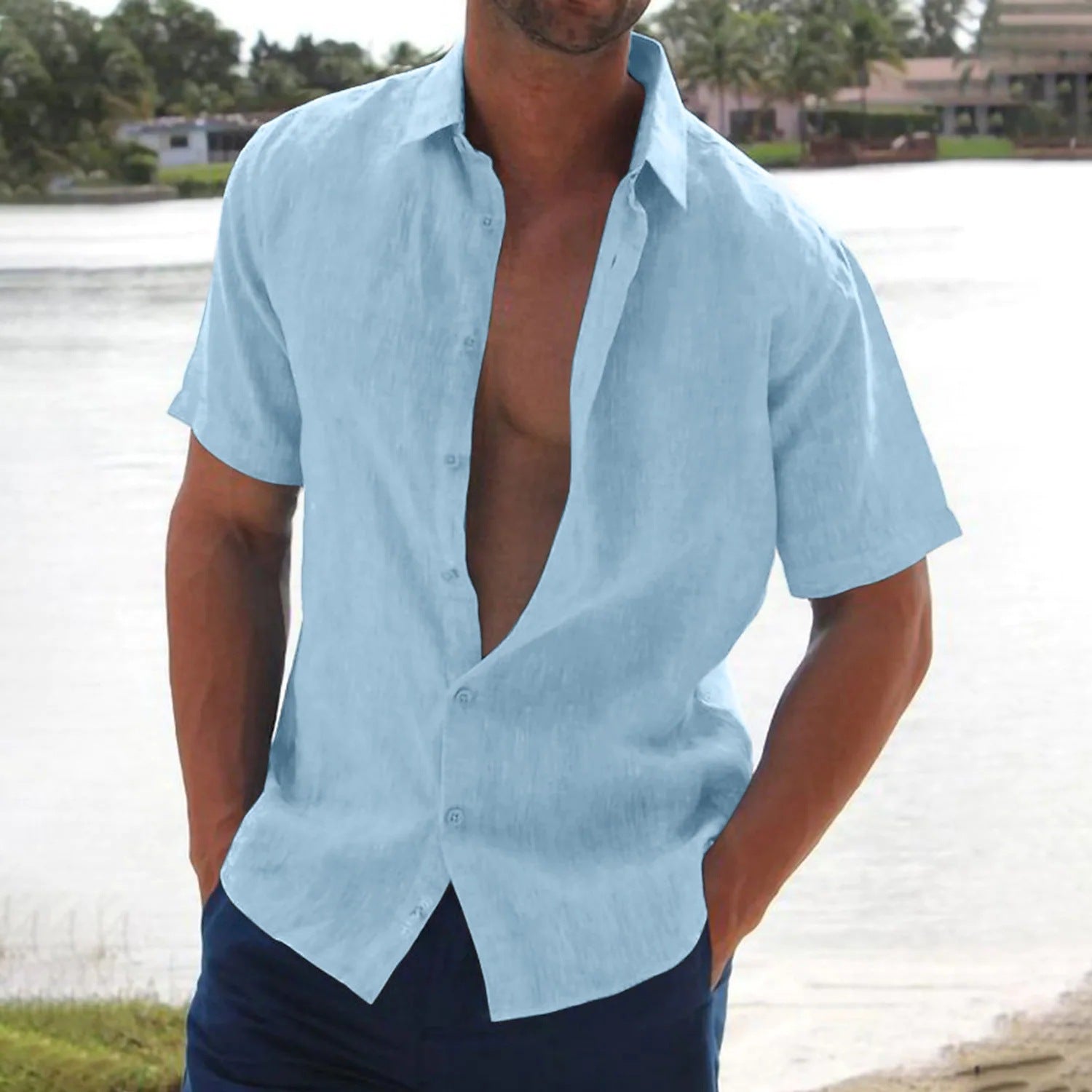 Man wearing a light blue button-up shirt with the top buttons undone, standing by a body of water.