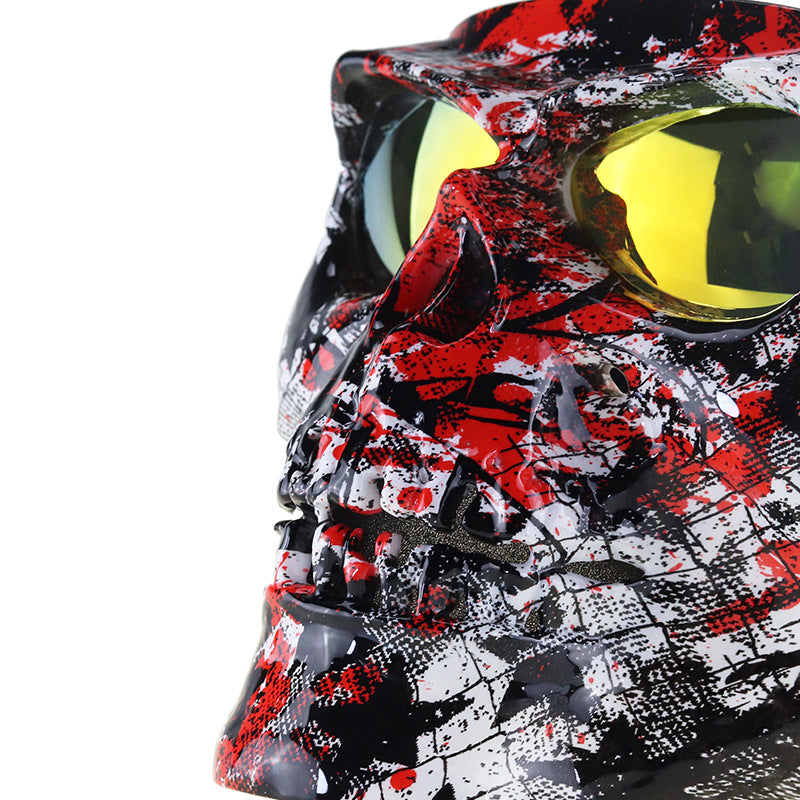 A close-up side view of a skull-shaped protective face mask or visor with a red, black, and white splatter pattern and reflective yellow lenses.